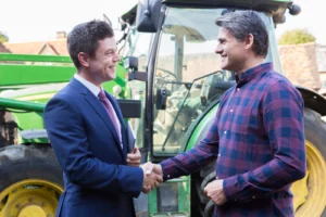 Farmer And Businessman Shaking Hands With Tractor In Background